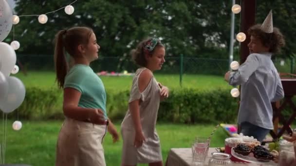 Two girls and a boy in a birthday hat have a fan around the set table. Cheerful teenagers dancing at a birthday party. Childrens birthday, catering concept — Stock Video