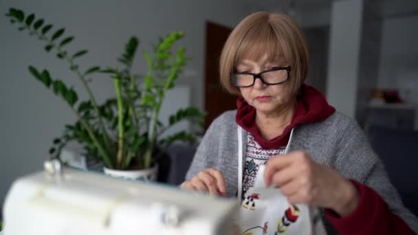 Mujer usando máquina de coser en casa. Mujer mayor cose textiles para el hogar utilizando la máquina. Madre hace ropa usando habilidades de costura, tela e hilo — Vídeos de Stock