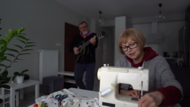 Et eldre par blir hjemme sammen. En pensjonist syr på en skrivemaskin. Mannen hennes spiller gitar. Lykkelig par hjemme under lockdown, Valentines dag – stockvideo