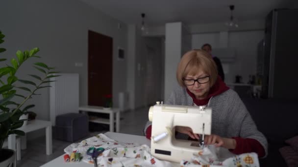 Una coppia felice di pensionati a casa loro. La moglie scarabocchi su una macchina da cucire, il marito le porta il tè con limone. Rimanere a casa concetto, lavorare a casa — Video Stock