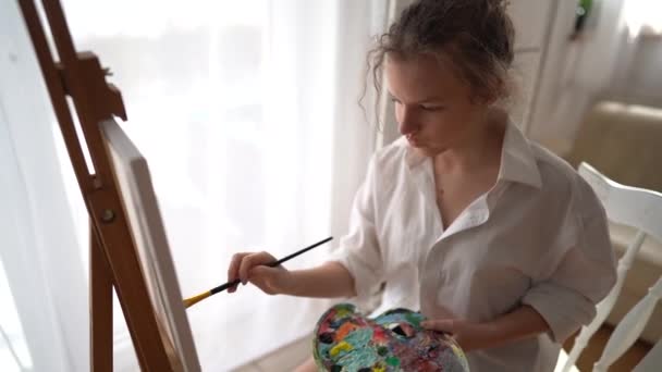 Portré egy gyönyörű tinédzser lányról fehér ingben, festményen ecsettel a vásznon. Egy fiatal művész ül egy széken egy állvány mellett, és rajzol egy képet — Stock videók