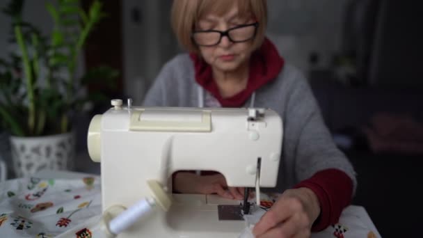 Зрелая женщина в очках шьет домашний текстиль на швейной машинке. Пенсионерка и ее хобби, бабушка работает на дому — стоковое видео