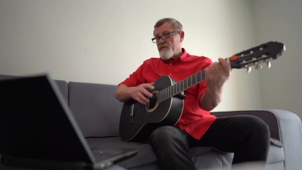 Пожилой человек или музыкант в очках и красной рубашке с ноутбуком компьютер учится играть на гитаре с учителем онлайн дома. Концепция музыки, электронного обучения и технологий — стоковое видео