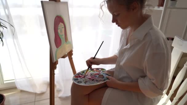 Detailní záběr jemné dívky v bílé košili malování portrét na plátně. Dívka sedí na židli u stojanu a v rukou drží paletu a štětec. Malířství a hobby koncept — Stock video