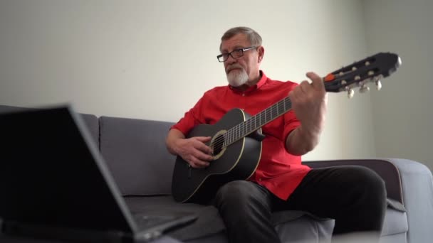 Gözlüklü ve kırmızı balıklı yaşlı gri sakallı adam evinde kanepede otururken dizüstü bilgisayar kullanarak video dersi veriyor. — Stok video