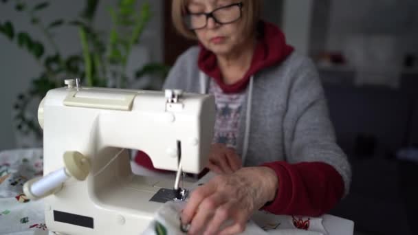 Blízký portrét starší švadleny. Důchodce šije záclony na šicím stroji doma — Stock video