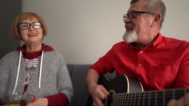 El marido toca la guitarra y la esposa canta la canción. Retrato de una pareja de ancianos que tiene un ventilador en su sofá en casa. Quédate en casa concepto — Vídeos de Stock