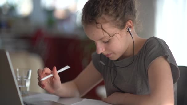 Dzieci, edukacja i koncepcja odległego uczenia się - nastoletnia studentka w słuchawkach z laptopem w domu. Cute girl słucha uważnie, odpowiedzi i pisze w notatniku — Wideo stockowe