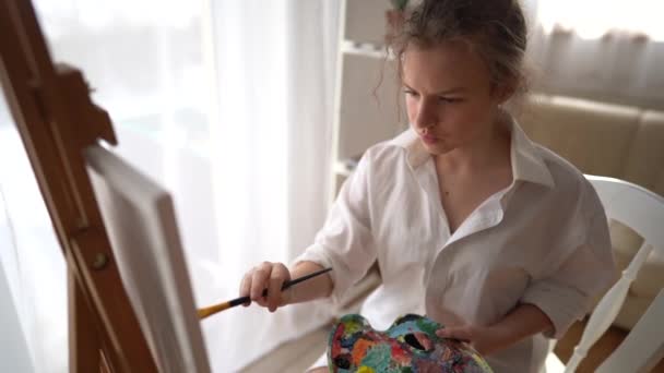 Menina bonita em uma camisa branca com escova na mão senta-se perto de um cavalete e desenha na tela. Criativa adolescente menina paitning uma imagem no cavalete. Menina praticando desenho na escola de arte — Vídeo de Stock