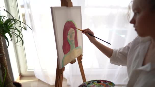 Dívka sedí na židli u stojanu a v rukou drží paletu a štětec. Malířský a hobby koncept. Detailní záběr jemné dívky v bílé košili malování portrét na plátně — Stock video