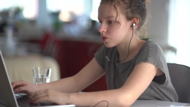 Gyermekek, oktatás és távoli tanulás koncepciója - tinédzser lány fülhallgatóban laptoppal otthon. Aranyos lány figyel, válaszol és ír egy jegyzetfüzetben — Stock videók