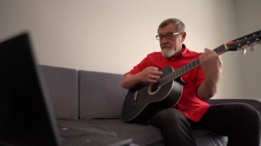 Olgun bir adam dizüstü bilgisayarın yanındaki koltukta otururken internetten gitar çalmayı öğrenir. Gözlüklü, evde gitar çalan gri sakallı bir emekli.