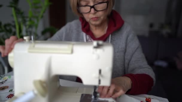 Пожилая швея во время работы и рабочий стол с ниткой, ножницами, иглами и измерительной лентой на нем — стоковое видео