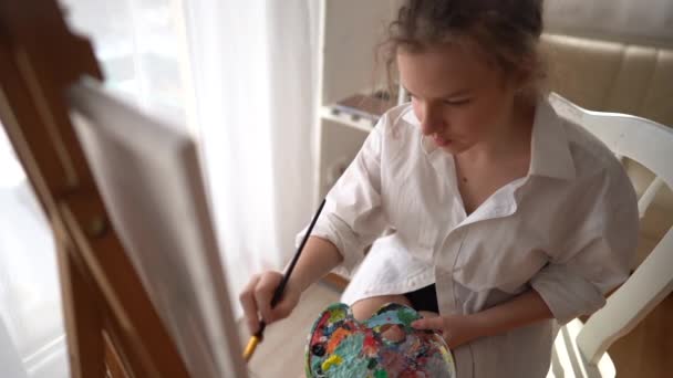 El primer plano de un joven artista se sienta en una silla cerca de un caballete y dibuja un cuadro. Retrato de una hermosa adolescente en una camisa blanca pintando con un pincel sobre lienzo. Vista superior — Vídeo de stock