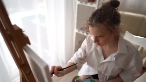 Chica joven pinta un retrato con pinturas al óleo en casa. La colegiala se sienta en una silla cerca del caballete y sostiene una paleta y un cepillo en sus manos. Niños ocio y creatividad — Vídeo de stock