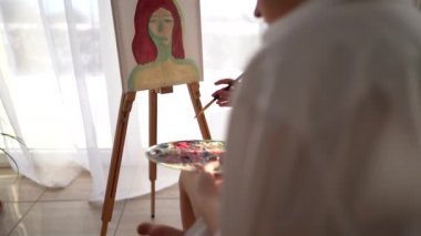 Kız sehpanın yanındaki sandalyeye oturuyor ve elinde palet ve fırça tutuyor. Resim ve hobi konsepti. Beyaz tişörtlü kibar bir kızın tuvale bir portre çizmesine yaklaş.