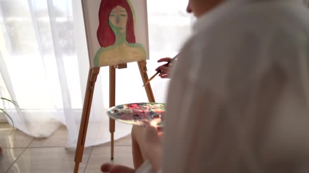 Dívka sedí na židli u stojanu a v rukou drží paletu a štětec. Malířský a hobby koncept. Detailní záběr jemné dívky v bílé košili malování portrét na plátně — Stock video