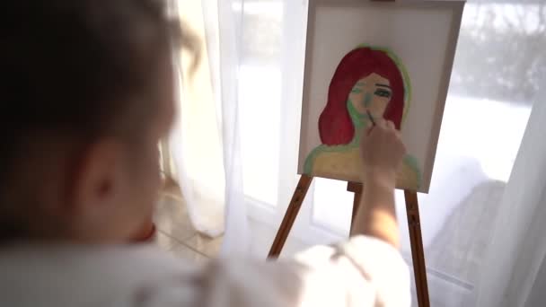 Zadní pohled. Krásný portrét mladé dívky v bílé košili malující portrét na plátně. Roztomilý umělec sedí u stojanu a maluje štětcem, zatímco sedí doma u okna — Stock video