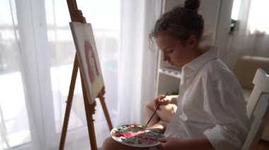 Kız sehpanın yanındaki sandalyeye oturuyor ve elinde palet ve fırça tutuyor. Resim ve hobi konsepti. Beyaz tişörtlü kibar bir kızın tuvale bir portre çizmesine yaklaş.