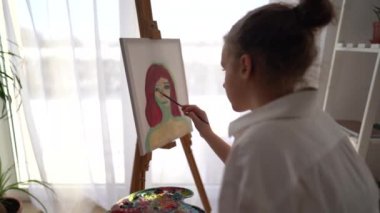 Kız sehpanın yanındaki sandalyeye oturuyor ve elinde palet ve fırça tutuyor. Resim ve hobi konsepti. Beyaz tişörtlü kibar bir kızın tuvale bir portre çizmesine yaklaş.