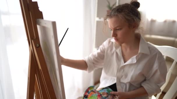 Chica en una camisa blanca pinta un cuadro con pinturas al óleo. Una adolescente se sienta cerca de la ventana con un caballete y pinta con un pincel sobre lienzo. Concepto de ocio y hobby para niños — Vídeo de stock