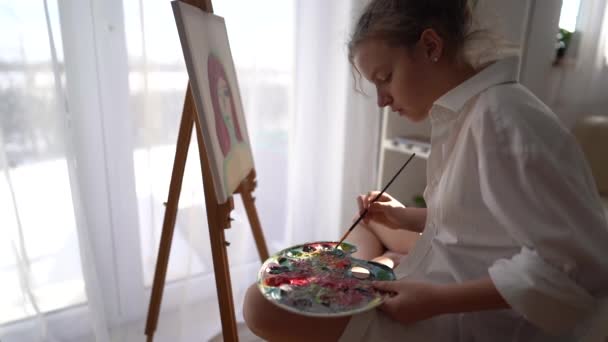 女性のティーンエイジャーは、帆布にブラシでイーゼルと塗料で窓の近くに座っています。子供のレジャーや趣味の概念。白いシャツの少女は油絵具で絵を描く — ストック動画