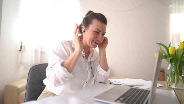 Gadis manis dengan kemeja putih memiliki panggilan video di kantor. Wanita tertawa dan berbicara melalui panggilan video menggunakan laptop dan headphone — Stok Video