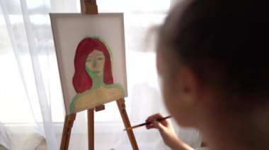 Güzel genç bir sanatçı, yağlı boyalarla bir portre çiziyor. Evinde pencerenin önünde oturuyor. Evde kal konsepti