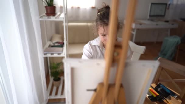 La colegiala aprende a pintar con pinturas al óleo. Una chica se sienta frente a un caballete y un lienzo y sostiene un pincel y una paleta con pinturas en las manos — Vídeo de stock