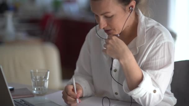 Uma mulher de negócios séria tem uma chamada de vídeo no escritório. Lady em uma camisa branca usa um laptop e fones de ouvido com fio, fala enquanto segura um microfone, faz anotações em um notebook — Vídeo de Stock