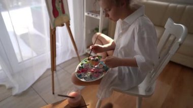 Kız sehpanın yanındaki sandalyeye oturuyor ve elinde palet ve fırça tutuyor. Resim ve hobi konsepti. Beyaz tişörtlü kibar bir kızın tuvale bir portre çizmesine yaklaş.