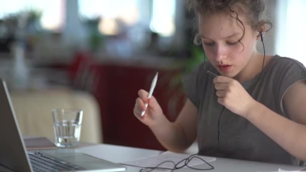 Portré egy iskolás lányról, aki laptopot használ, miközben online tanul. A lány bedrótozott fülhallgatóból beszél a mikrofonba, és leírja a választ egy jegyzetfüzetbe. — Stock videók