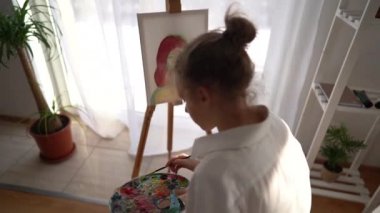 Kız sehpanın yanındaki sandalyeye oturuyor ve elinde palet ve fırça tutuyor. Resim ve hobi konsepti. Beyaz tişörtlü kibar bir kızın tuvale bir portre çizmesine yaklaş.