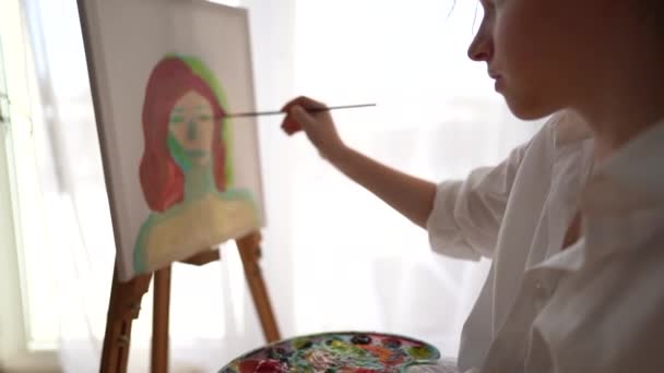 Portred of the girl painting a picture. Schoolgirl learns to draw. Teenage girl draws a self-portrait while sitting on a chair near the window — Stock Video
