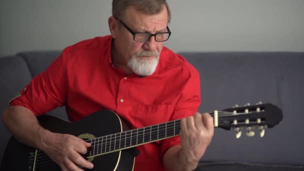 Senior man playing acoustic guitar at home. Portrait of a gray-haired mature man in a red shirt and glasses playing the guitar while sitting on the couch at home. Stay home concept — Stock Video