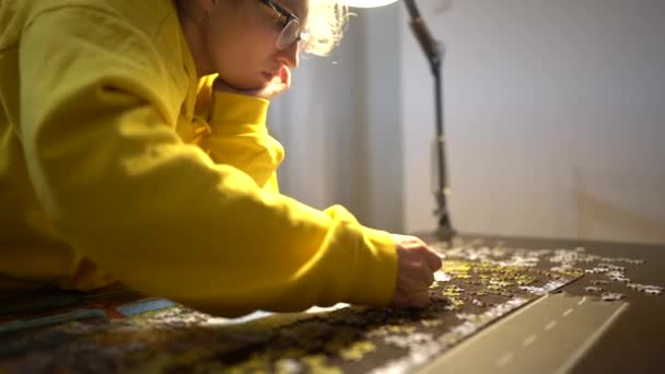 Femme portant des lunettes et haut jaune faisant puzzle à la maison sous la lumière d'une lampe de table. Femme assise au bureau à la maison et résoudre un puzzle, concept de loisirs et de jeux, rester à la maison — Video