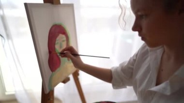 Kız sehpanın yanındaki sandalyeye oturuyor ve elinde palet ve fırça tutuyor. Resim ve hobi konsepti. Beyaz tişörtlü kibar bir kızın tuvale bir portre çizmesine yaklaş.