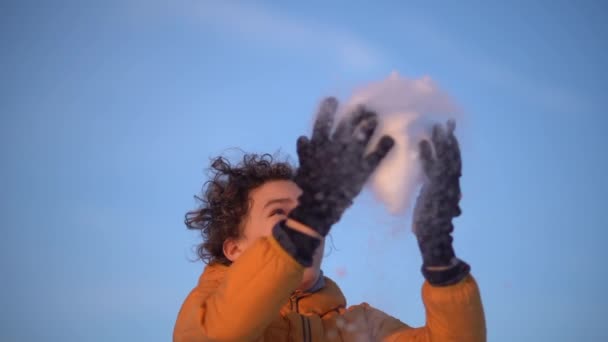 Portret van een vrolijke gekrulde schooljongen die sneeuw gooit. De jongen draagt een geel jasje en zwarte leren handschoenen — Stockvideo