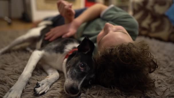 Кудрявый школьник и его собака лежат на ковре в помещении. Мальчик и собака, домашние животные и дети — стоковое видео