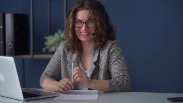 Empleo remoto durante el coronavirus. Jovencita en el cargo. Mujer de negocios sonriente con auriculares que utilizan el ordenador portátil durante la videollamada — Vídeos de Stock