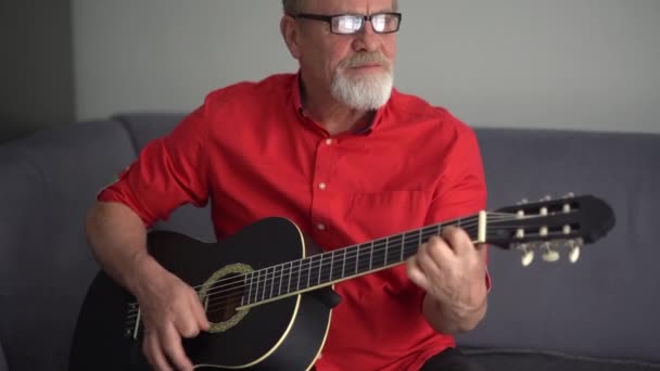 Retrato de um pensionista tocando uma guitarra interior. O homem está tocando ativamente, pegando acordes, separando os trastes — Vídeo de Stock
