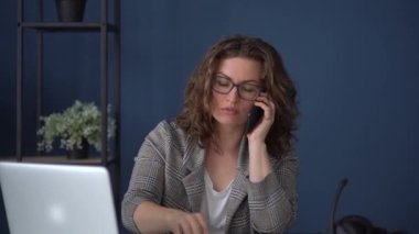 Ofisteki masasında birden fazla iş yapan bir kadının portresi. Kız telefonla konuşuyor, mikrofonlu kulaklık takıyor ve laptopla video bağlantısı kuruyor.