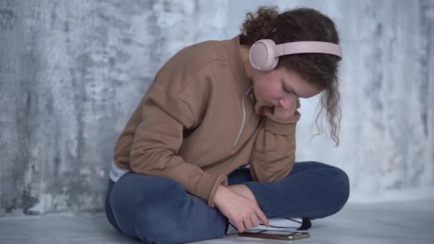 Menina adolescente triste senta-se sozinho perto de uma parede cinza, curvando-se sobre seu smartphone. A estudante usa auscultadores e ouve música. Pessoas e gadgets — Vídeo de Stock
