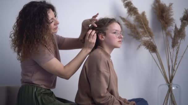 Schöne gelockte Frau webt ihre Teenager-Haare Zöpfe. Muttertag, Mutter und Tochter zu Hause. Glückliche Familie — Stockvideo