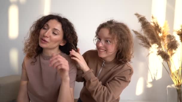 母亲和女儿在家里的肖像。少女把她可爱的成熟母亲的头发编成辫子 — 图库视频影像