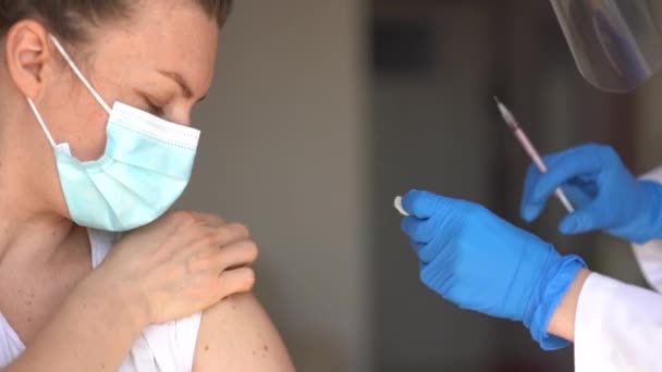 Inyección de cerca de una mujer joven con una máscara que recibe una vacuna contra el coronavirus en el hombro. Enfermera en máscara, escudo y guantes hace una inyección — Vídeos de Stock