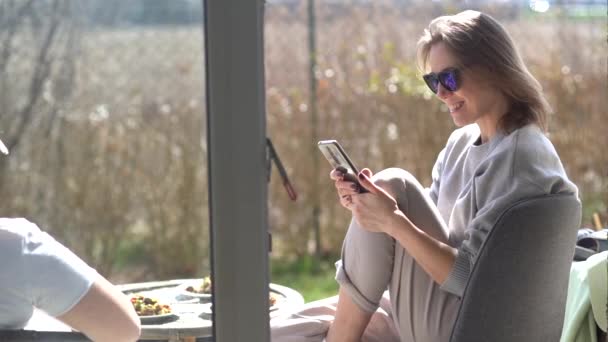 Amigos mujeres positivas viendo fotos en el teléfono inteligente mientras descansan en la terraza de primavera cerca de la casa. Concepto de ocio — Vídeos de Stock