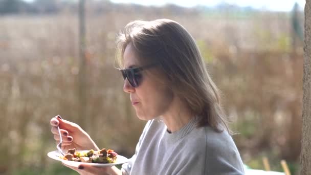 Dziewczyna w okularach zjada sałatkę z talerza i cieszy się pierwszymi promieniami wiosennego słońca. Piknik na tarasie wiejskiego domu — Wideo stockowe