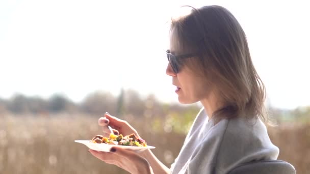 Chica con gafas de sol comiendo. Mujer madura con gafas come una ensalada de un plato y disfruta de los primeros rayos del sol de primavera. Picnic en la terraza de una casa rural — Vídeos de Stock