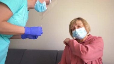 Yaşlılar için güvenli bağışıklık. Doktor yaşlı kadın hastaya grip aşısı yapıyor. Tıbbi maske takan olgun bir kadın evdeki koltukta oturuyor ve koluna Covid-19 aşısı yaptırıyor.